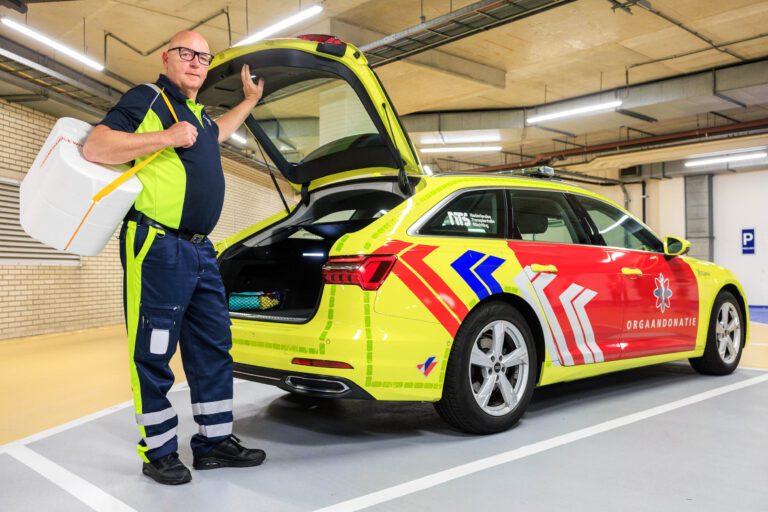 Dit is een foto van een man in een gele en blauwe uniform, klaar om een belangrijke taak uit te voeren. Hij staat naast een felgekleurde auto met opschrift Orgaandonatie in een parkeergarage. De kofferbak van de auto is open, en de man houdt een koelbox vast die waarschijnlijk bedoeld is voor het transport van organen. De auto is ook voorzien van reflecterende markeringen voor zichtbaarheid. De foto is genomen door Jan Buwalda in Groningen.