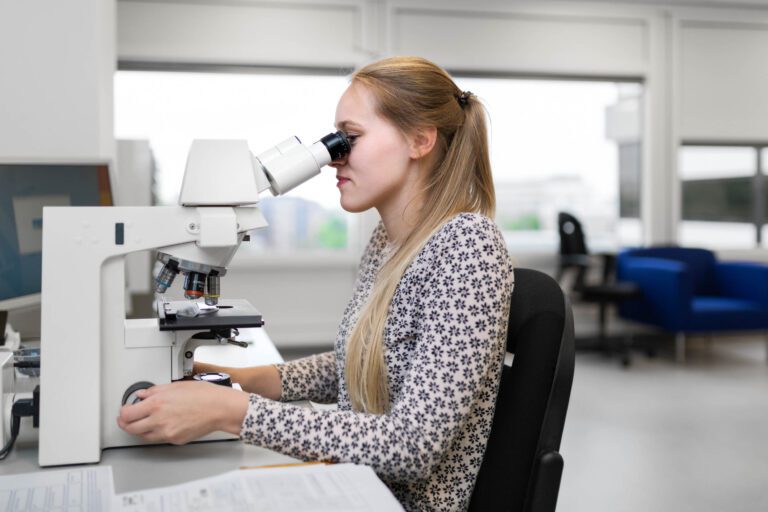 Een jonge vrouw met lang blond haar kijkt geconcentreerd door een microscoop in een moderne laboratoriumomgeving. Ze draagt een gebloemde blouse en zit op een bureaustoel. Op de achtergrond zijn onscherpe kantoorruimtes te zien. Foto door Jan Buwalda, genomen in Groningen.