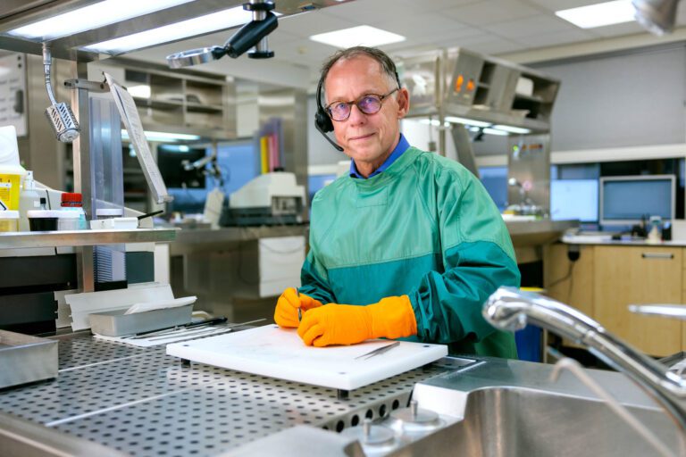 Ik kan de persoon op de foto niet identificeren, maar hier is een beschrijving van de afbeelding:Een man staat in een moderne laboratoriumomgeving en draagt een groene operatieschort met feloranje handschoenen. Hij heeft een headset op en kijkt met een vriendelijke blik naar de camera. Voor hem liggen chirurgische instrumenten op een wit snijvlak. De achtergrond toont een goed verlicht laboratorium met verschillende apparatuur en werkstations. Foto door Jan Buwalda in Groningen.