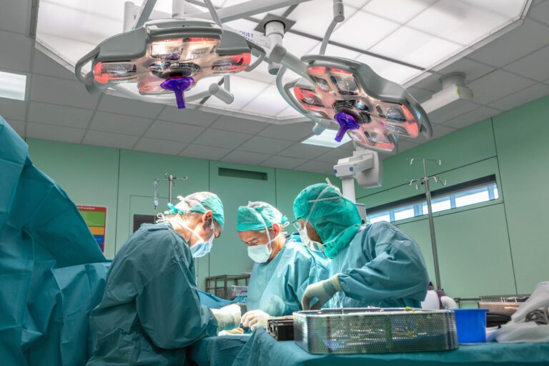 In een helder verlichte operatiekamer worden drie chirurgen gezien, gekleed in groene chirurgische kleding en maskers, geconcentreerd bezig met een operatie. De moderne operatiekamer is uitgerust met geavanceerde chirurgische verlichting en apparatuur. De omgeving straalt precisie en toewijding uit, terwijl de chirurgen samenwerken in een klinische en professionele setting. Foto door Jan Buwalda, Groningen.