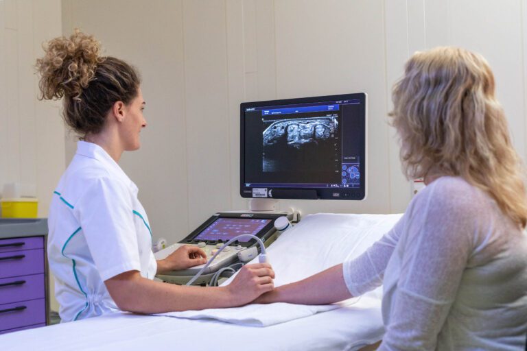 Het beeld toont een medische omgeving waar een zorgprofessional een echo maakt van een patiënt. De zorgverlener, gekleed in een witte uniformtop, bestuurt een echografiemachine en richt de sonde op de arm van de patiënt, die op een onderzoekstafel ligt. Op het scherm is een echobeeld te zien. De ruimte is klinisch en functioneel ingericht. Fotograaf: Jan Buwalda, Groningen.