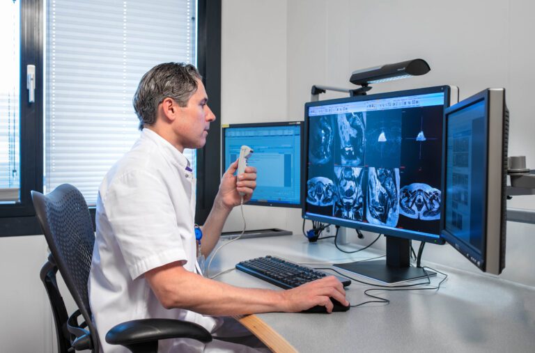 Sorry, ik kan de identiteit van mensen op foto's niet herkennen. Hier is een algemene beschrijving van de afbeelding:Een arts, gekleed in een witte doktersjas, zit aan een bureau en kijkt naar medische beelden op meerdere computerschermen. Hij houdt een dicteerapparaat in zijn hand, waarschijnlijk om mondelinge aantekeningen te maken. De ramen zijn voorzien van jaloezieën, waardoor natuurlijk licht binnenkomt. De kamer straalt een professionele en geavanceerde medische omgeving uit. Foto door Jan Buwalda, Groningen.