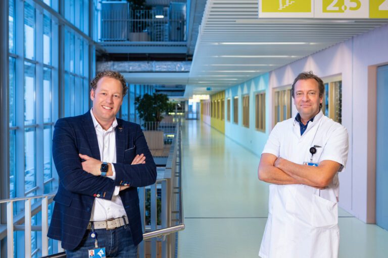 Het beeld toont twee mannen die ontspannen glimlachen en poseren met de armen over elkaar in een moderne, lichte gang van een gebouw. De man links draagt een donkerblauw pak met een licht overhemd en een polsbandje, terwijl de man rechts een wit doktersjasje draagt. De omgeving heeft grote glazen panelen met blauwe en gele tinten op de muren. De foto is genomen door Jan Buwalda in Groningen.