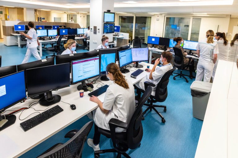 Dit is een levendige foto van een moderne ziekenhuisafdeling, vastgelegd door fotograaf Jan Buwalda in Groningen. Verschillende personeelsleden in witte uniformen zitten achter bureaus met meerdere computerschermen, terwijl anderen door de ruimte lopen. De blauwe vloer en heldere verlichting geven de omgeving een schone en georganiseerde uitstraling. Iedereen draagt een mondkapje, wat wijst op een medisch-professionele setting.