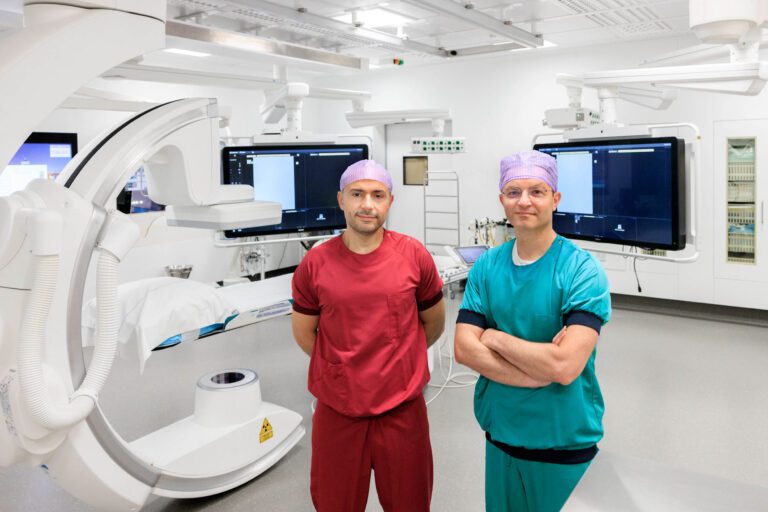 Twee medisch specialisten staan in een moderne operatiekamer, gehuld in gekleurde operatiekleding. De ruimte is uitgerust met geavanceerde medische apparatuur en grote beeldschermen. De sfeer is professioneel en steriel. Foto door Jan Buwalda, Groningen.