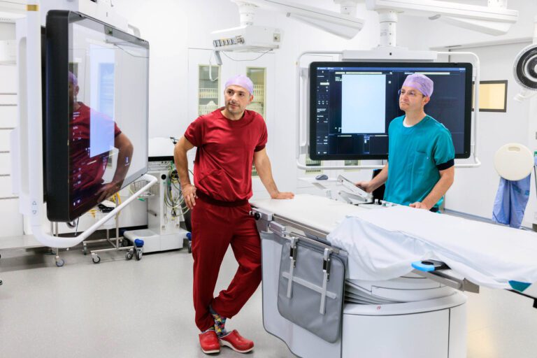 Dit beeld toont een moderne medische onderzoeksruimte met twee medische professionals. De persoon links draagt een rood uniform en paarse operatiemuts en leunt ontspannen tegen een console. De ander, rechts, draagt een groen uniform en observeert een monitor. Om hen heen zijn geavanceerde medische apparatuur en schermen te zien. Fotograaf: Jan Buwalda, Groningen.