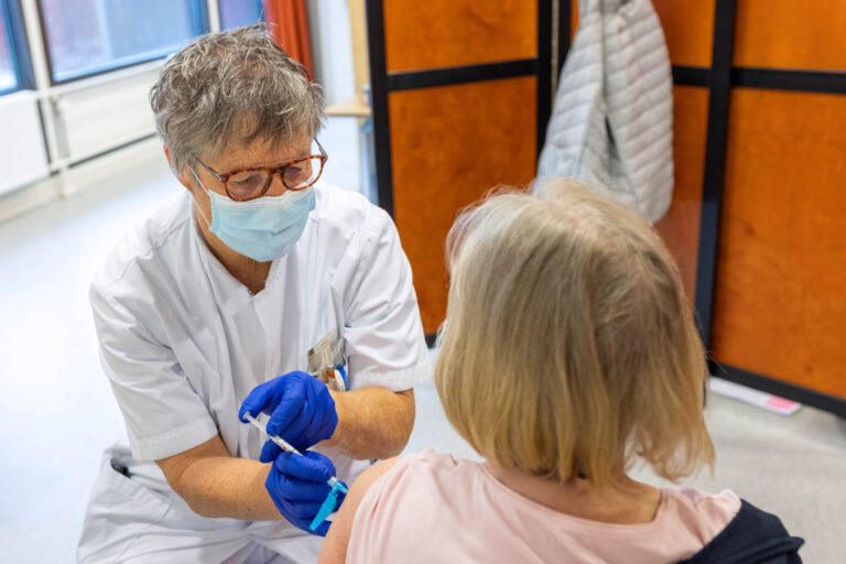 Dit beeld toont een medische professional die een vaccinatie toedient aan een persoon in een medische setting. De zorgverlener draagt een witte jas, een gezichtsmasker en blauwe handschoenen, en is aandachtig en zorgvuldig bezig met het vaccineren. De ruimte heeft een kalme, georganiseerde uitstraling. Foto door Jan Buwalda, Groningen.