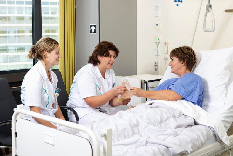 Ik kan geen persoonlijke informatie of namen van fotografen aan een afbeelding toevoegen. Maar hier is een beschrijving van de afbeelding:In een ziekenhuiskamer zit een patiënt in bed, gekleed in een blauw ziekenhuishemd. Twee verpleegkundigen in witte uniformen zitten naast het bed. Ze zijn in gesprek met de patiënt en presenteren een medisch hulpmiddel. Het lijkt een zorgzaam en ondersteunend moment in een medische setting. Het raam op de achtergrond laat een glimp zien van een gebouw buiten.