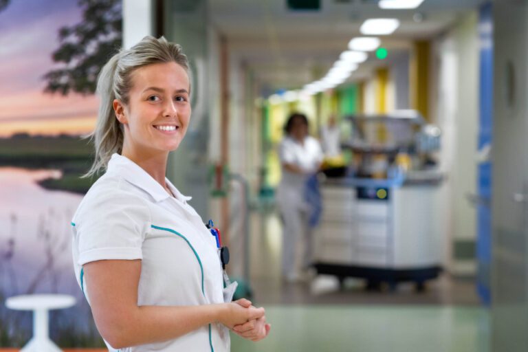 Een verpleegkundige staat met een vriendelijke glimlach in een heldere en moderne ziekenhuisgang. Op de achtergrond is een collega bezig met het verplaatsen van een karretje. De verlichting en het schilderij van een landschap aan de muur dragen bij aan een aangename sfeer. Foto door Jan Buwalda, Groningen.