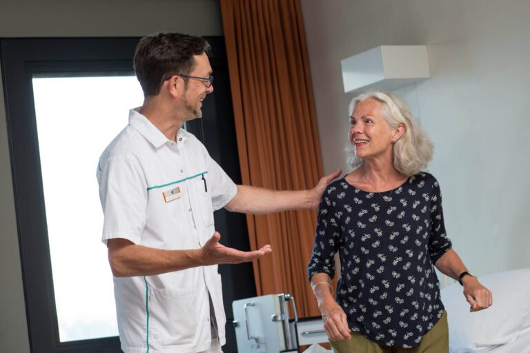 Dit is een foto van een medische professional die glimlachend met een vrouw praat in een kamer. De vrouw lijkt ontspannen en tevreden terwijl ze met hem communiceert. De man draagt een uniform dat typisch is voor medische professionals, terwijl de vrouw casual is gekleed. Ze staan samen bij een bed, wat suggereert dat ze zich in een zorgomgeving bevinden. Fotograaf: Jan Buwalda, Groningen.