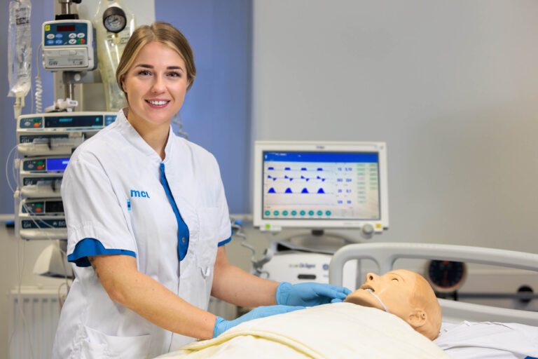 Ik kan geen namen van mensen in foto's herkennen. Maar ik kan wel een beschrijving maken.Op deze foto is een verpleegkundige te zien die glimlachend in een medische omgeving staat. Ze draagt een wit uniform met blauwe accenten. Haar handen zijn gehuld in blauwe handschoenen en rusten op de borst van een medische oefenpop die op een ziekenhuisbed ligt. Op de achtergrond is er medische apparatuur te zien, waaronder een monitor met grafieken en gegevens.Foto door Jan Buwalda, Groningen.