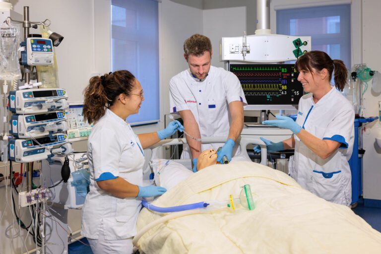 Dit is een foto van medisch personeel dat werkt in een ziekenhuisomgeving. Drie zorgverleners in witte uniformen en blauwe handschoenen zijn bezig met een medische pop in een bed, vermoedelijk voor trainingsdoeleinden. Op de achtergrond zijn diverse medische apparaten en monitoren zichtbaar, die vitale functies weergeven. De kamer heeft een neutrale uitstraling met blauwe en witte elementen. Foto door Jan Buwalda, Groningen.