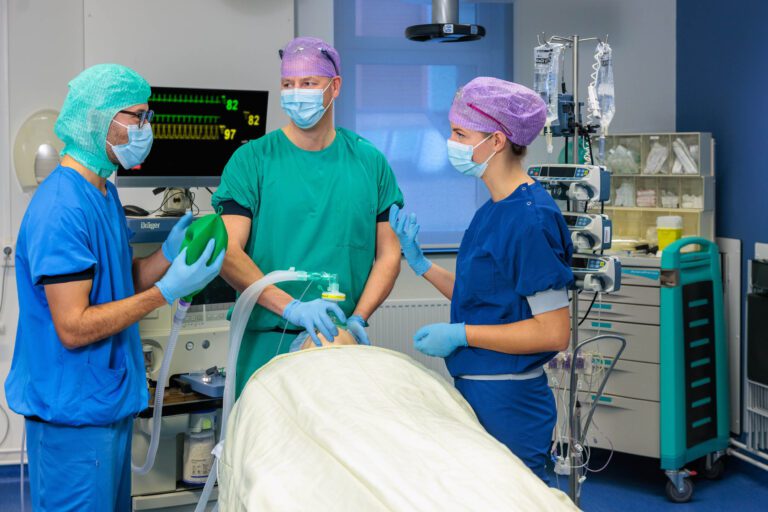 Dit beeld toont een medisch team in een operatiekamer dat bezig is met de voorbereiding van een operatie. Drie zorgverleners, gekleed in chirurgische scrubs en maskers, staan rond een operatiebed waarop een patiënt ligt bedekt met een laken. Een van hen houdt een beademingsballon vast, terwijl de anderen communiceren en zich concentreren op hun taken. Op de achtergrond zijn medische apparatuur en infuusstandaarden zichtbaar. Fotografie door Jan Buwalda, genomen in Groningen.