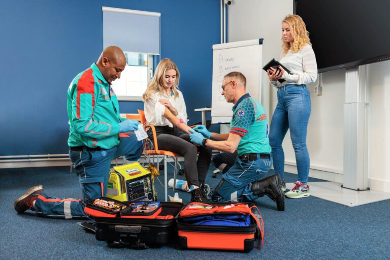Dit beeld toont een medische trainingssessie in een leslokaal. Twee hulpverleners in uniform, uitgerust met handschoenen, zijn bezig met een simulatie op een jonge vrouw die op een stoel zit. Ze gebruiken diverse medische apparatuur die uitgestald is op de grond. Op de achtergrond staat een vrouw met een tablet en een whiteboard met de tekst A, B, C, D in grote letters. Deze foto is genomen door Jan Buwalda in Groningen.