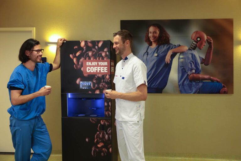 Twee mannen in medische kleding staan bij een koffieautomaat en kletsen met elkaar. De man in het blauw draagt een bril en leunt ontspannen tegen de automaat met een kopje in zijn hand. De andere man draagt een wit uniform en heeft eveneens een kop koffie vast. Achter hen hangt een poster met een afbeelding van een vrouw in blauwe scrubs die naast een crash test dummy zit. Het licht is warm en zacht, en de sfeer lijkt vriendelijk en ontspannen. Fotograaf: Jan Buwalda.